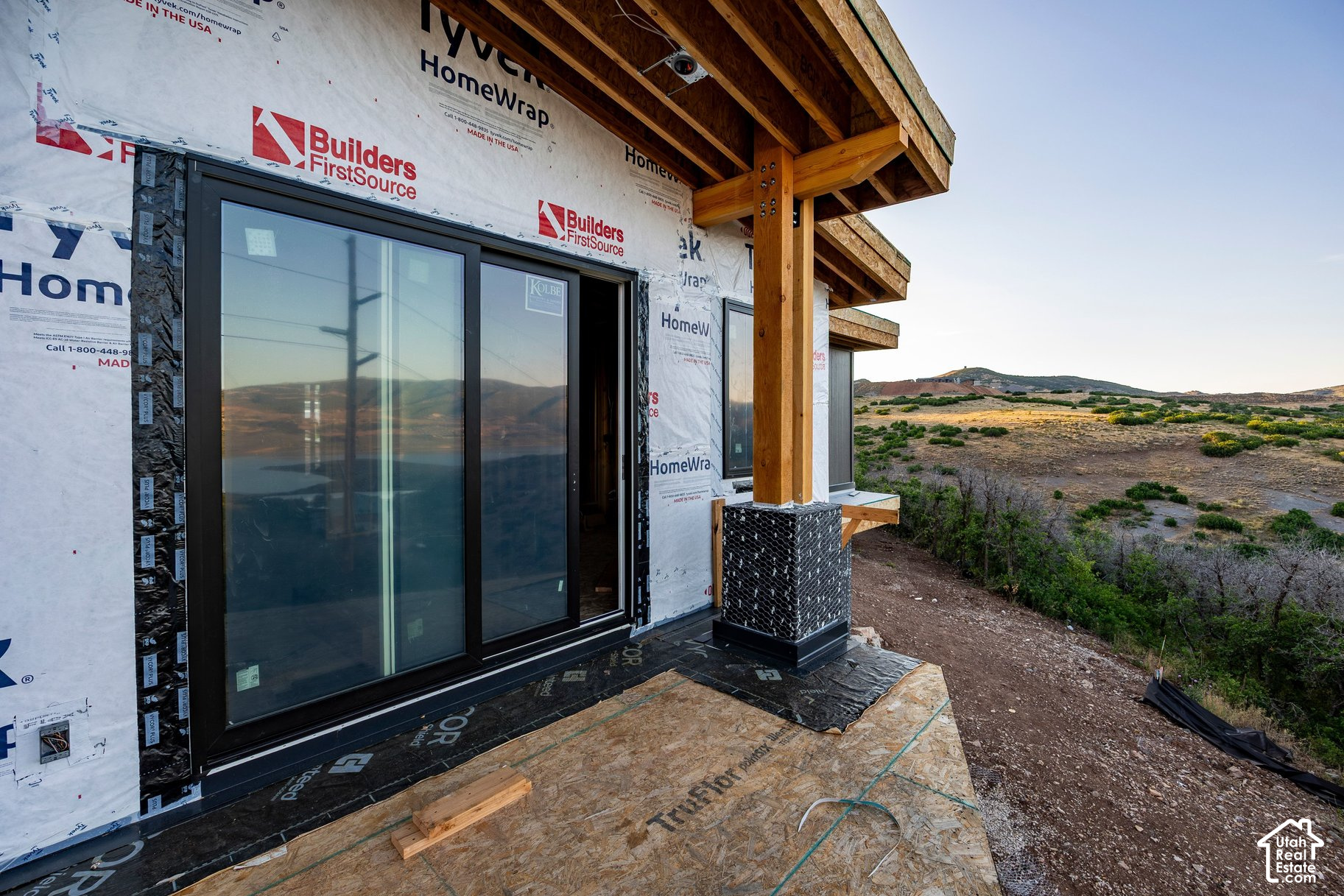 House under construction with a timber-framed porch and sliding glass door beside shrub-covered hills.