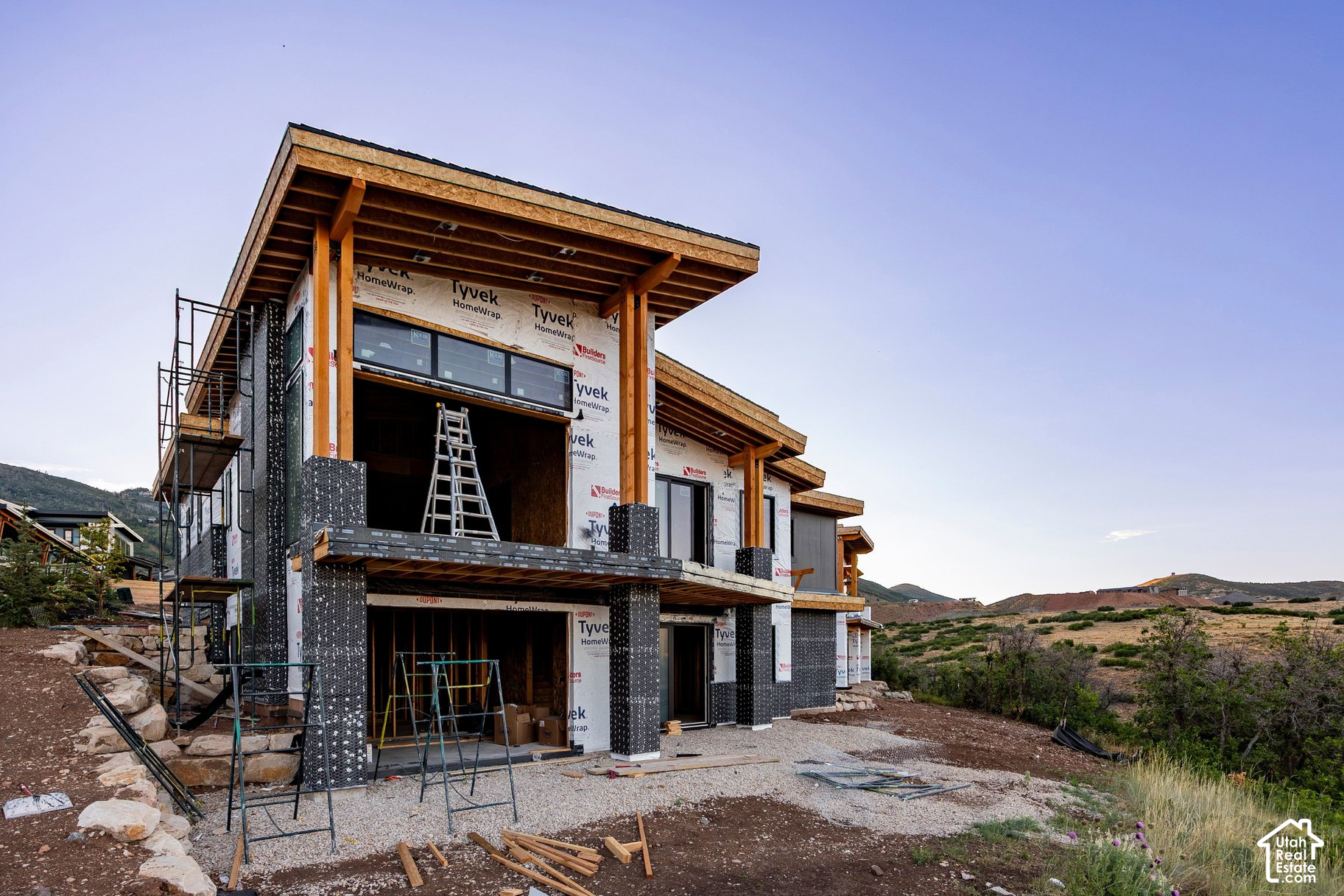 Modern two-story home under construction, with exposed framing and scaffolding against a hilly landscape.