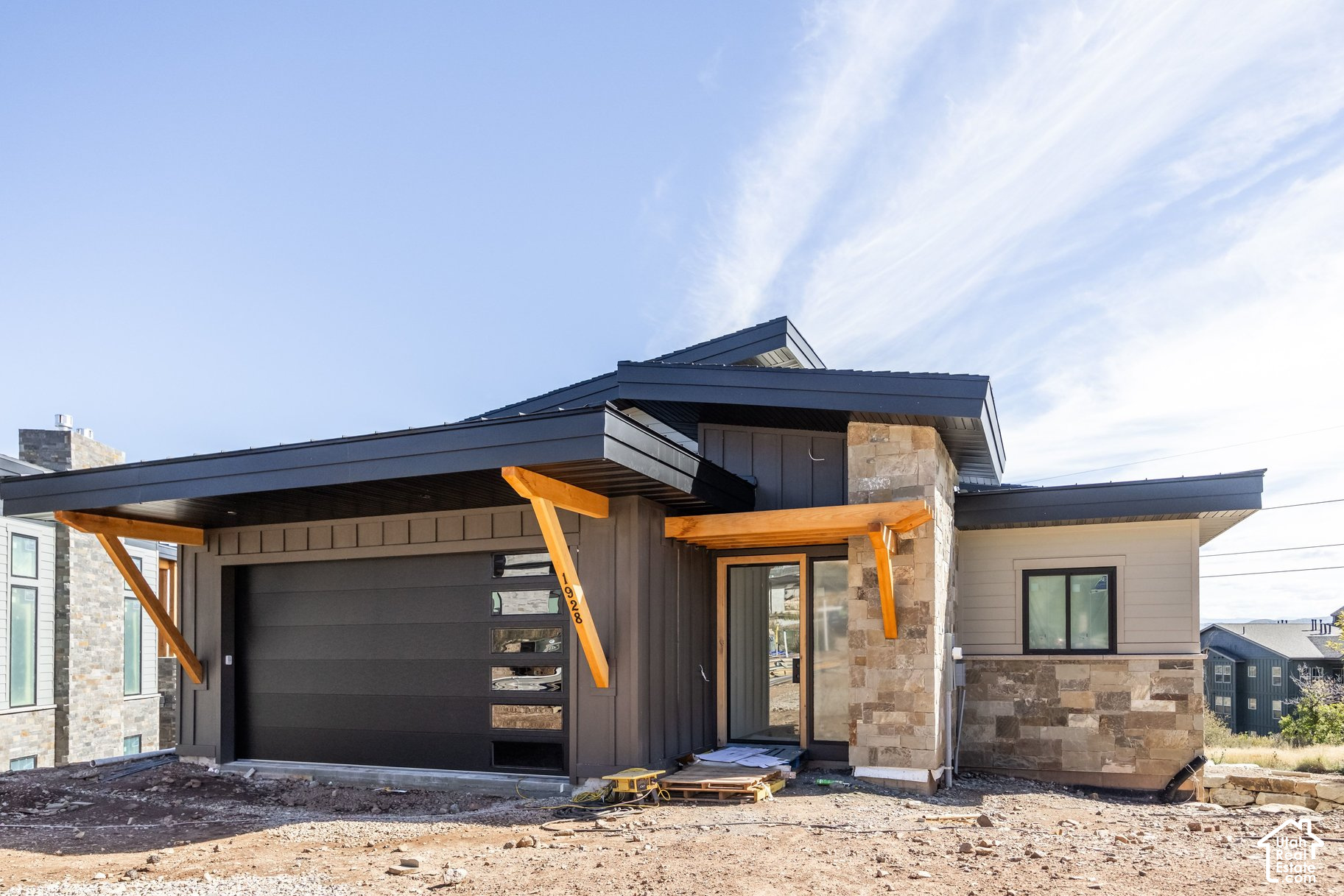 Modern home under construction with dark siding, stone facade, and timber supports over the garage.