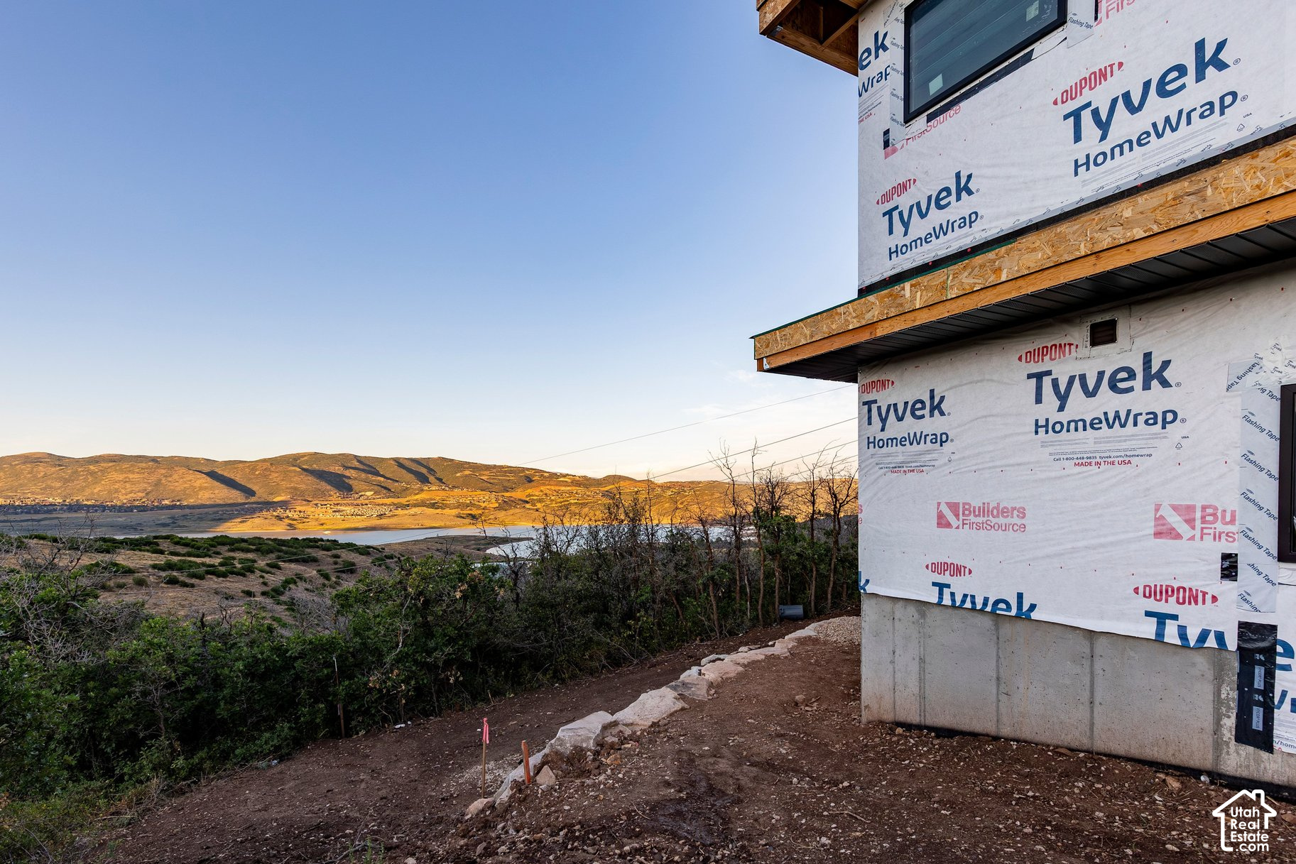 House under construction overlooking a lake and sunlit hills, with Tyvek-wrapped walls and exposed foundation.