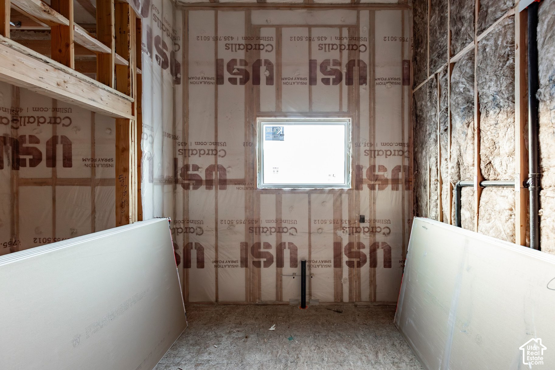 Unfinished room with exposed insulation and plumbing, a window, and drywall panels leaning against the walls.