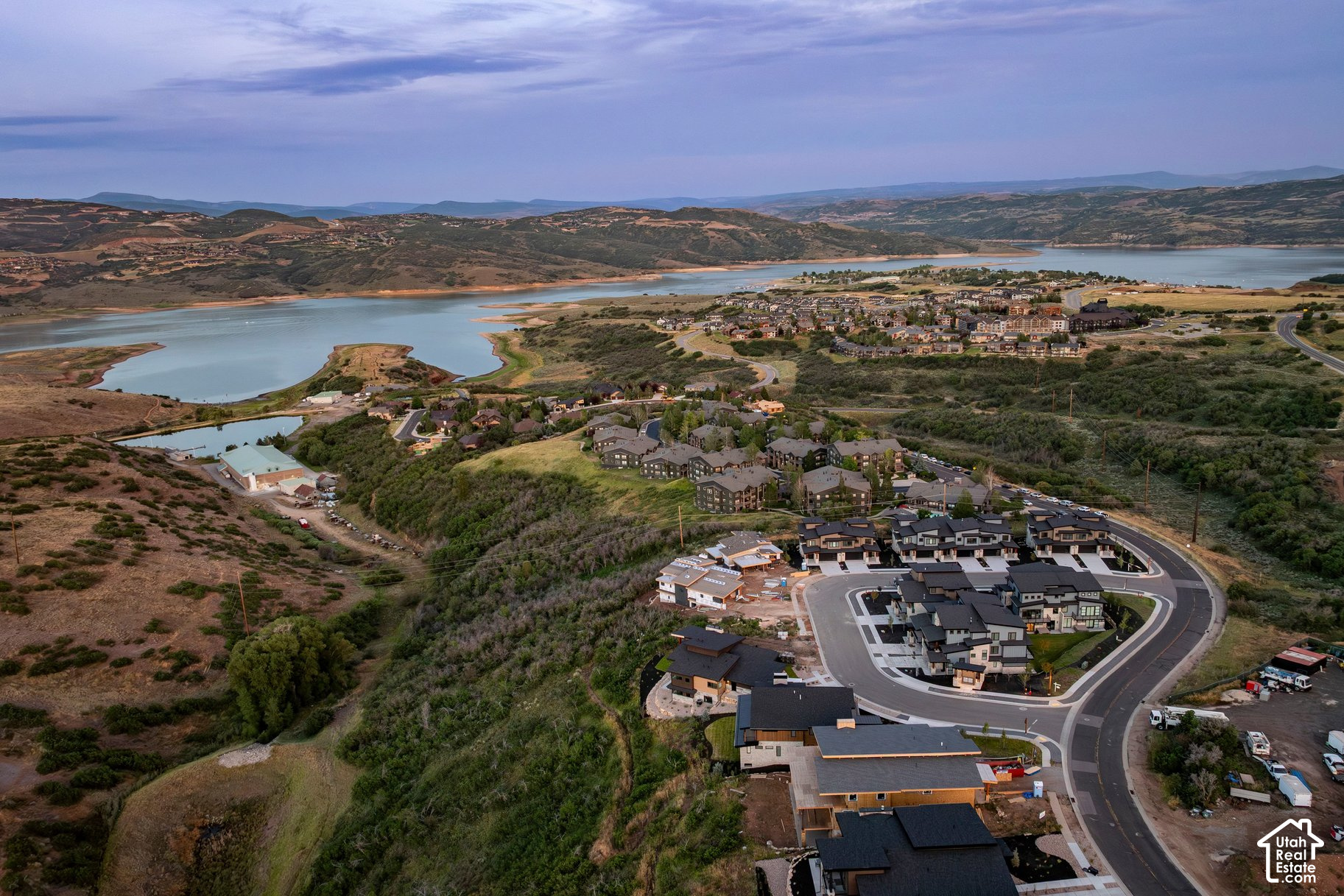 Aerial view of homes under construction beside a reservoir, with rolling hills and mountains beyond.