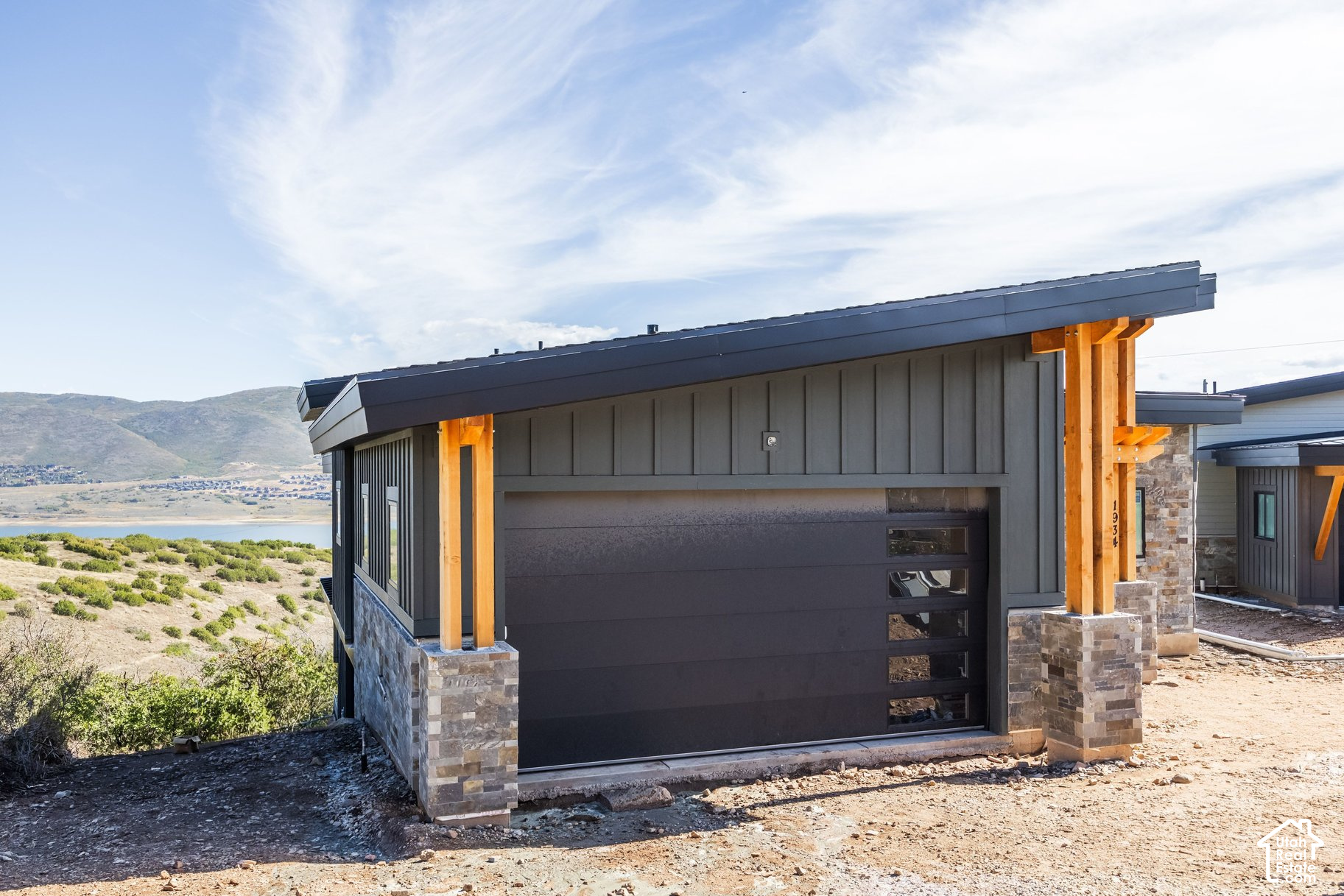 Modern dark-gray home with timber supports and stone base overlooking a lake and mountains.