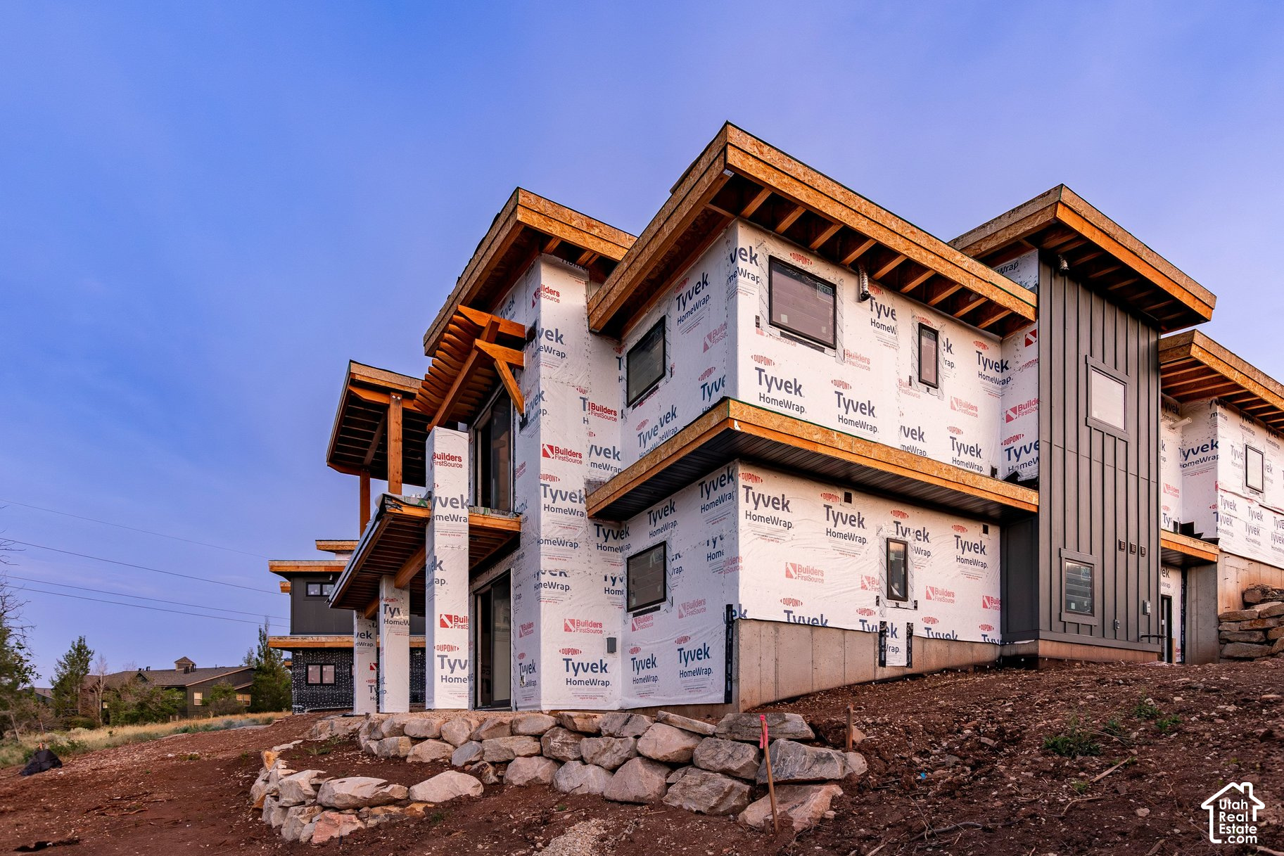 Modern multi-story home under construction with Tyvek wrap, exposed roof framing, and a rocky yard.