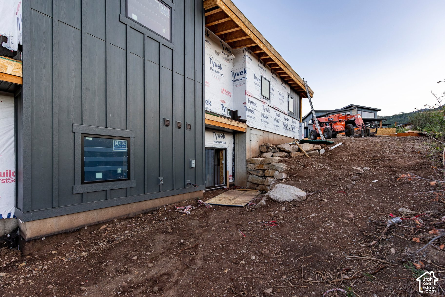 Modern home under construction with dark siding, exposed house wrap, and a lift on rocky ground.