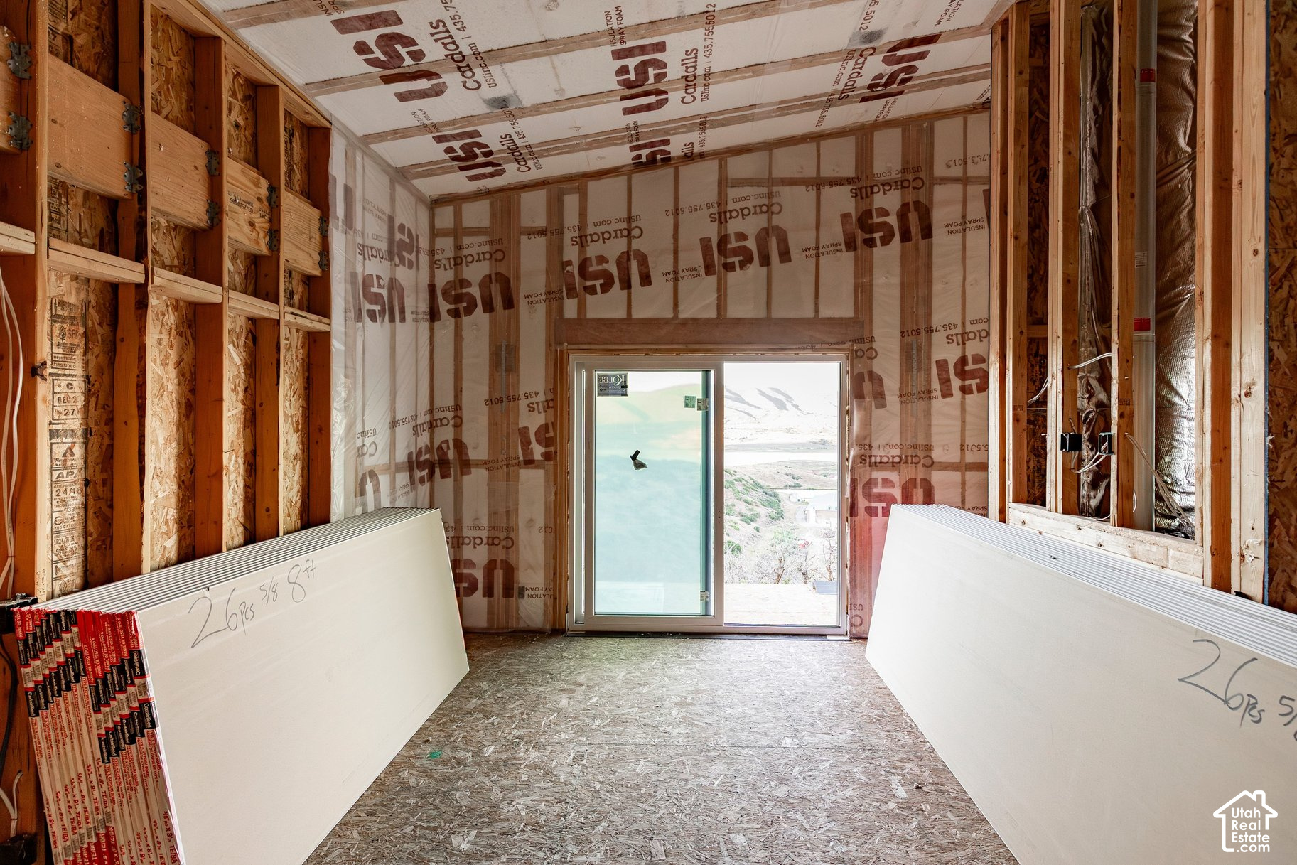 Unfinished framed room with insulation and stacked wallboard beside a sliding glass door overlooking water and snowy mountains.