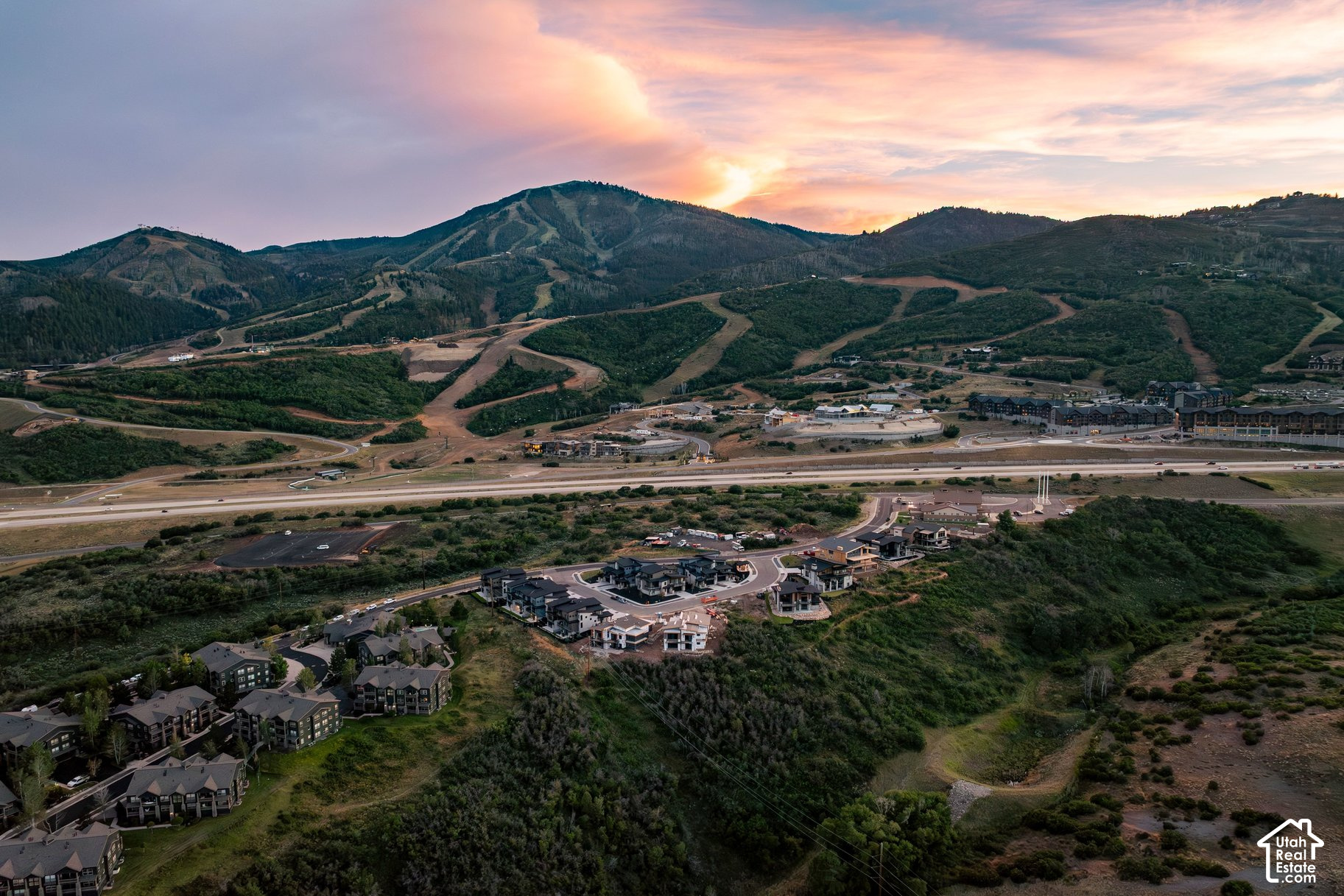 Aerial view of a mountain development at dusk, with homes and wooded hills beneath a pink sky.