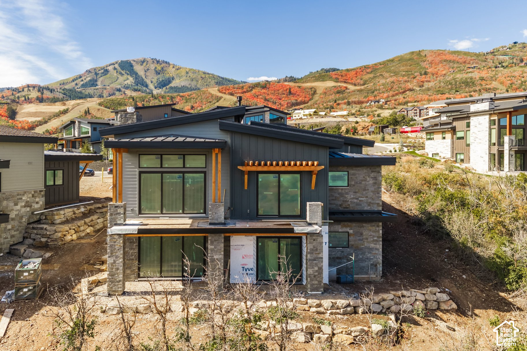 Modern mountain home under construction, with dark siding, stone accents, and autumn hills behind it.