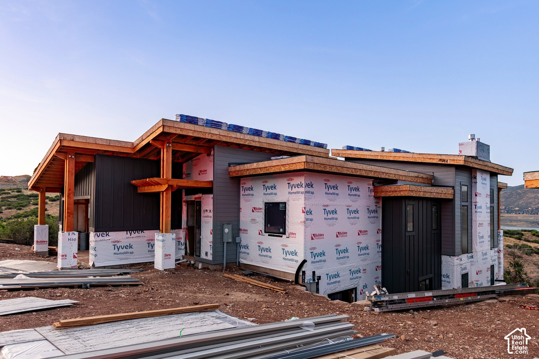 Modern home under construction with exposed timber beams and Tyvek-wrapped walls, with water and hills in the distance.