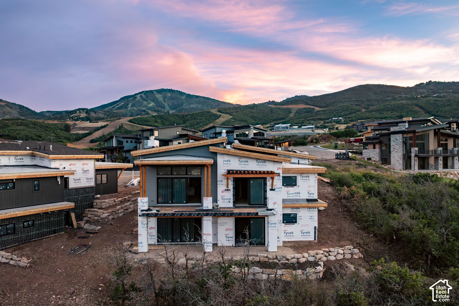 Modern home under construction with exposed house wrap, hillside homes, and mountains at dusk.