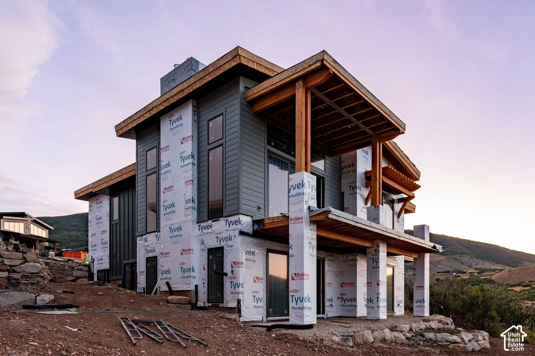 Modern hillside home under construction with exposed Tyvek wrap and prominent timber roof beams.