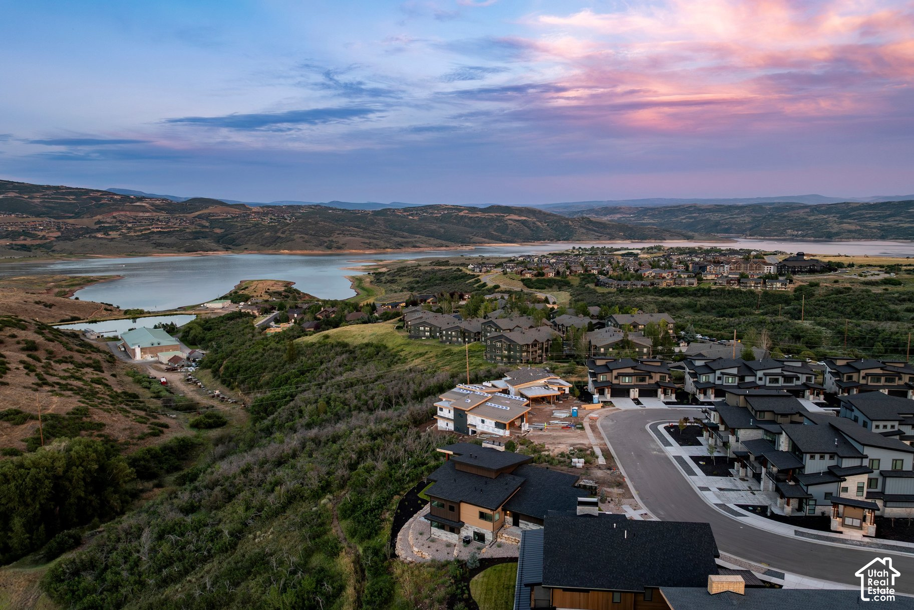 Aerial view of a hillside neighborhood, lake, and surrounding hills beneath a pink dusk sky.