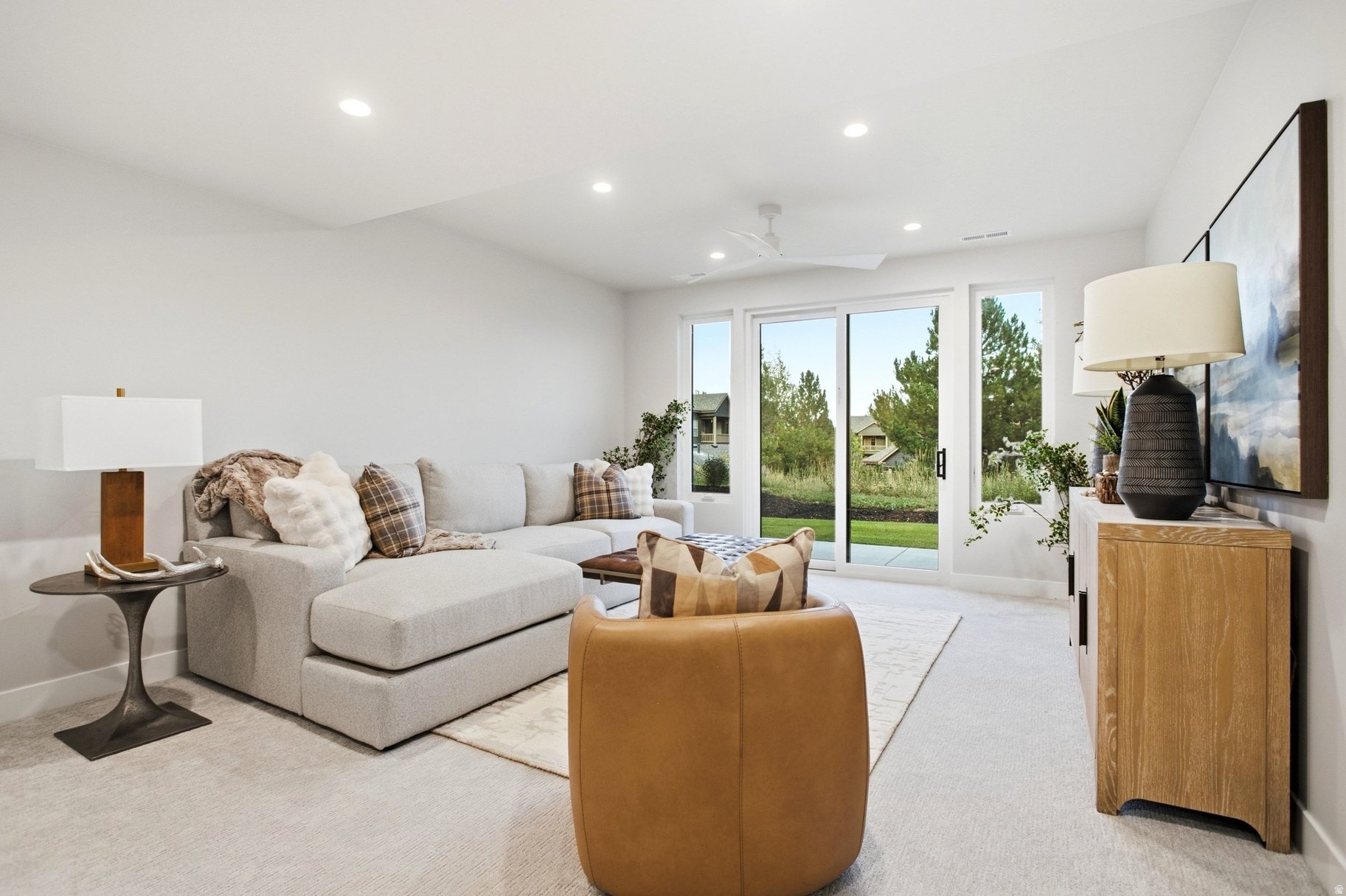 Staged living room with a gray sectional, tan leather chair, and wide glass doors overlooking greenery.