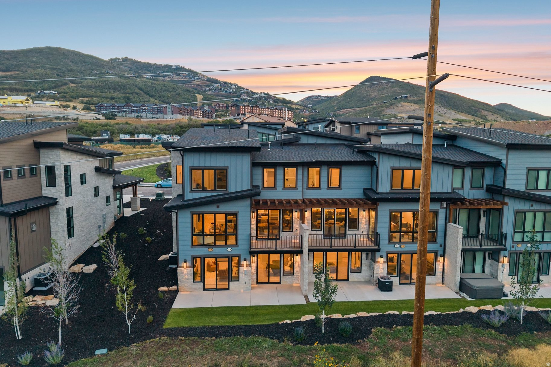 Modern townhomes with warm window lights sit below rolling hills at sunset.