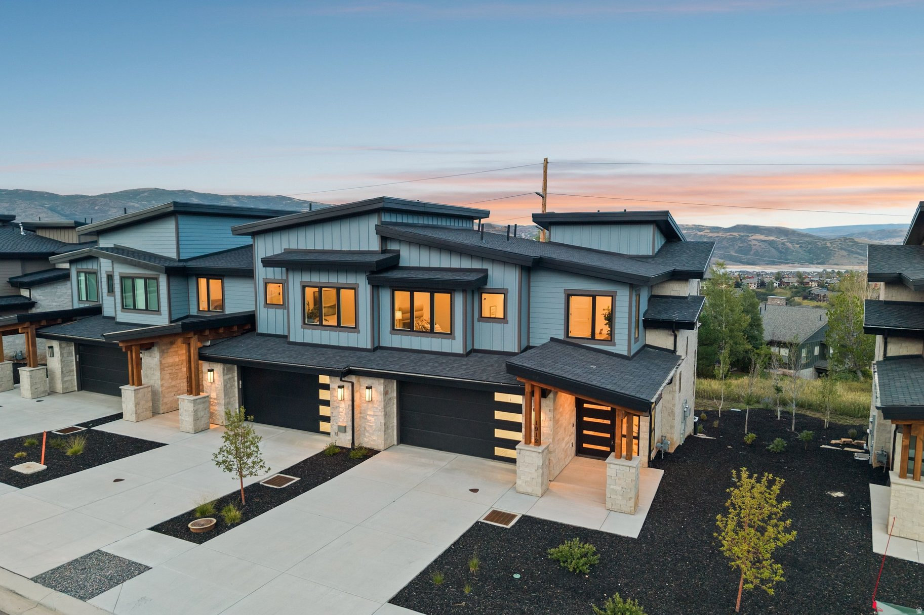 Modern blue-gray home with stone accents and lit windows, backed by mountain ridges at dusk.