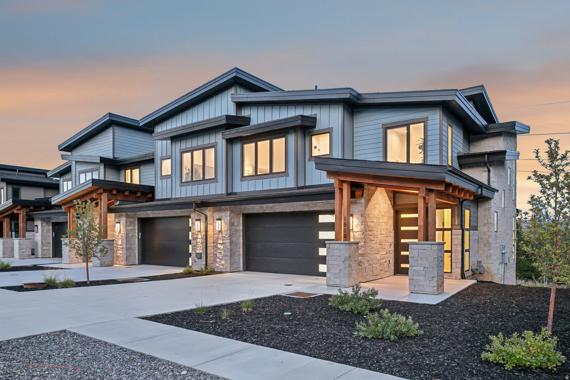 Modern two-story home with blue-gray siding, stone veneer, and a timber-covered entry at dusk.