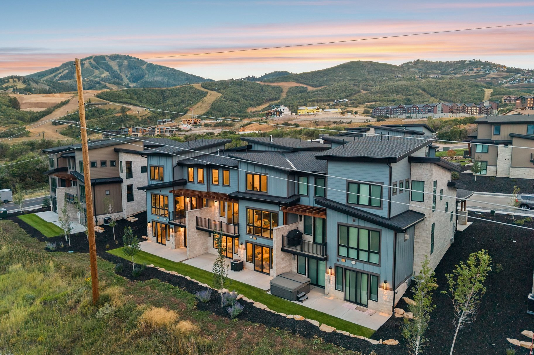 Modern gray-and-stone homes overlook a landscaped neighborhood and wooded mountain hills at sunset.