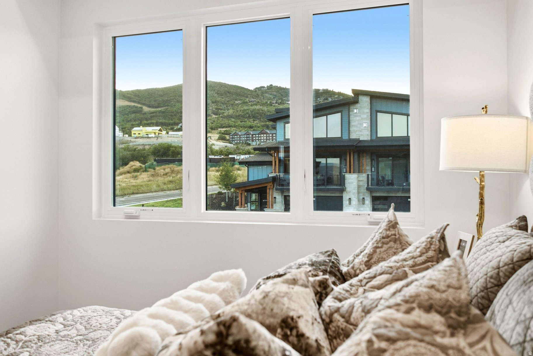 Bedroom with patterned pillows and wide windows overlooking a neighboring home and green hills.