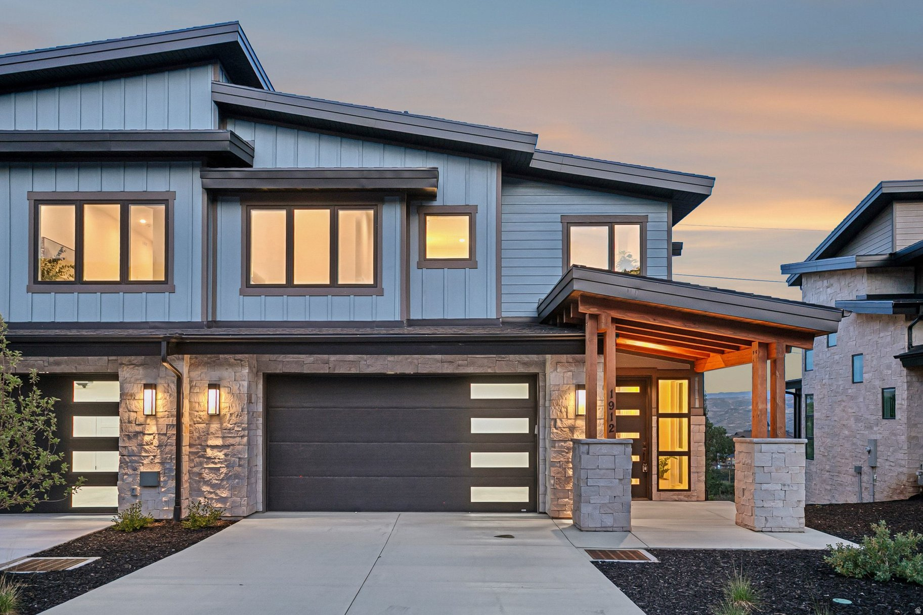Modern two-story home with stone facade, wide garage, and warmly lit covered entry at dusk.