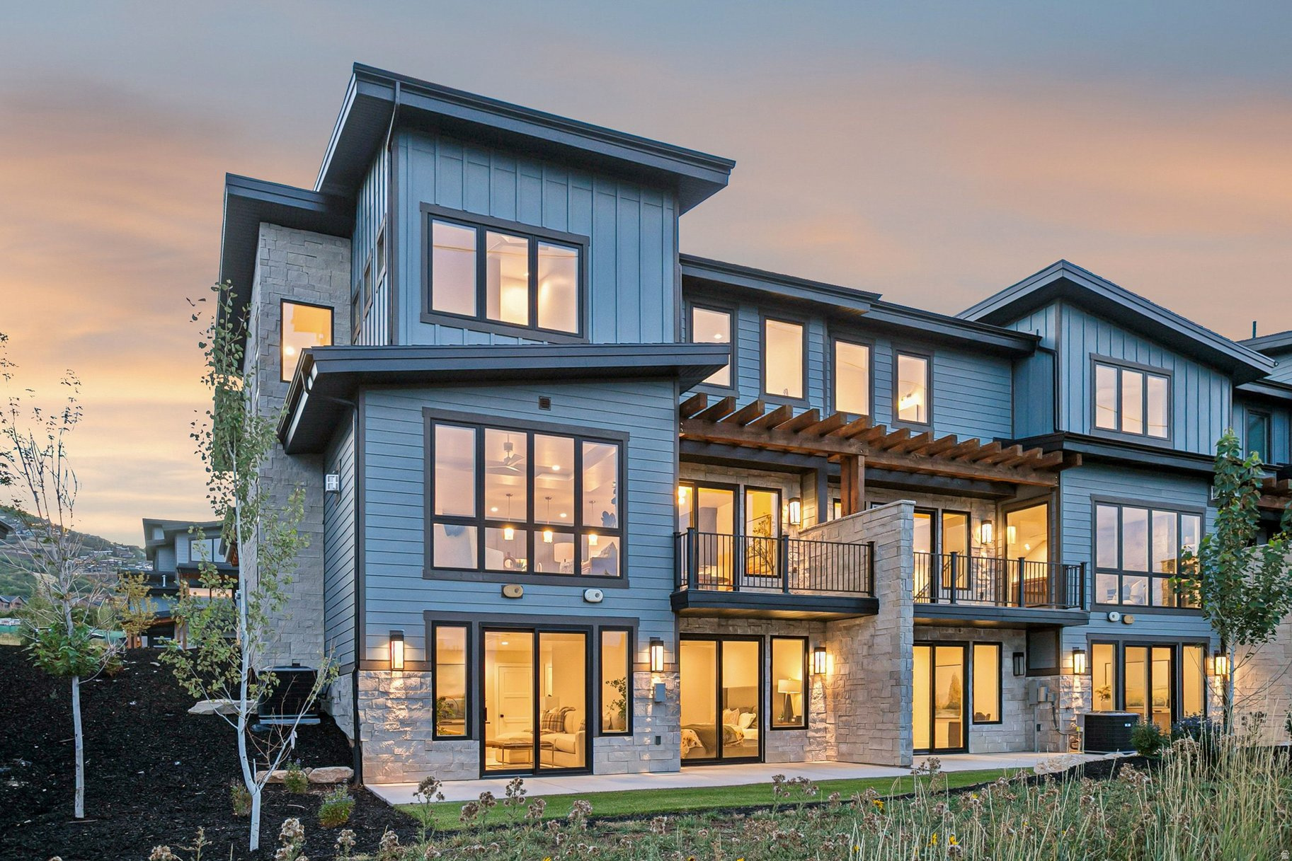 Modern blue-gray home with stone accents, glowing windows, balconies, and a timber pergola at dusk.