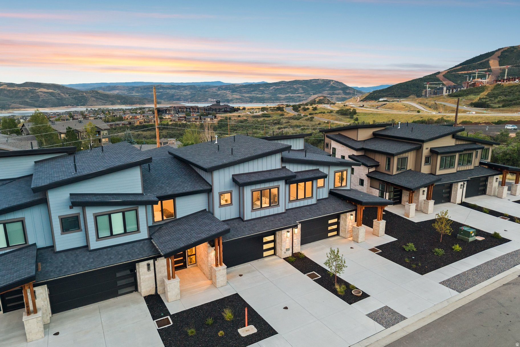 Modern homes with illuminated windows overlook a lake and mountains at sunset.