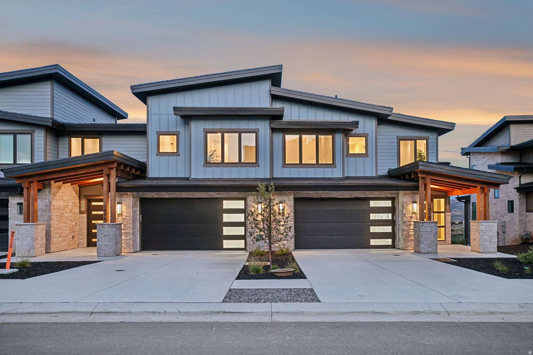 Modern two-story home with stone walls, twin garages, and covered timber entries at dusk.