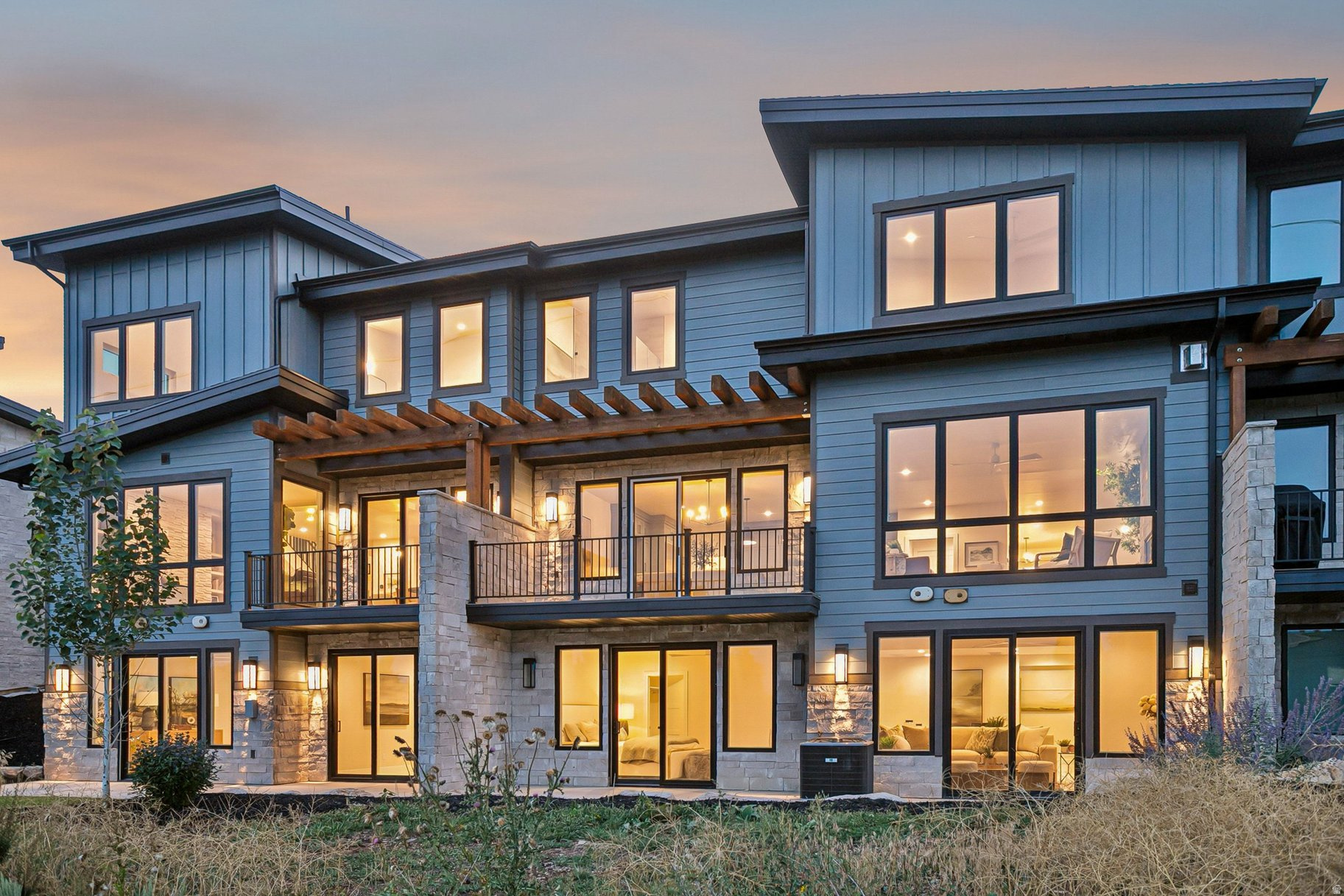 Contemporary multi-story home with stone accents, timber pergola, and warmly lit windows at dusk.