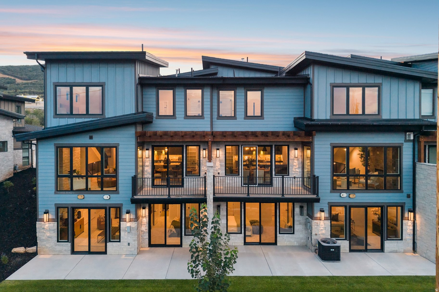 Modern blue-sided home with stone accents and a central balcony, glowing with warm interior lights at dusk.