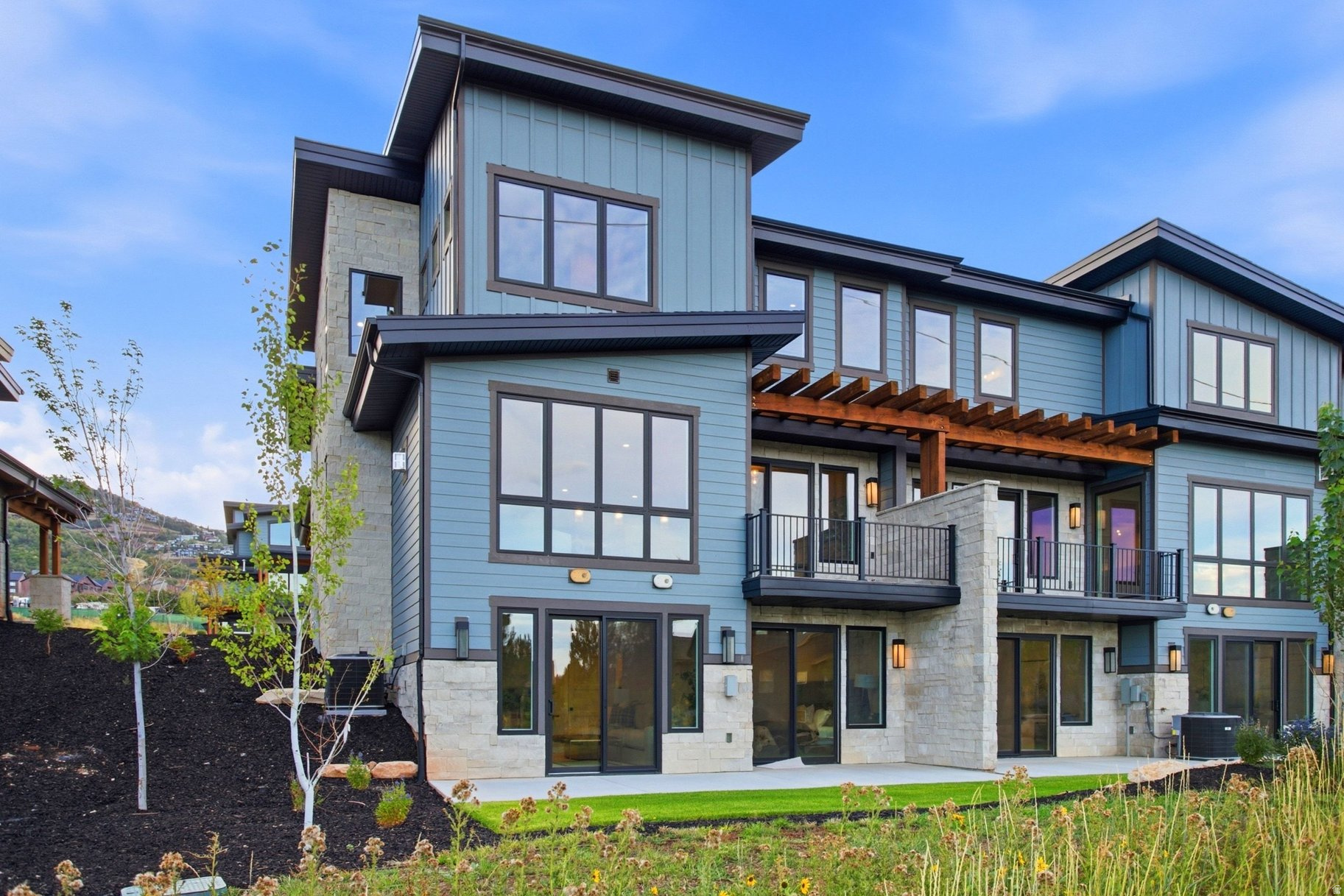 Modern blue-gray home with stone accents, balconies, and a timber pergola on a landscaped slope.