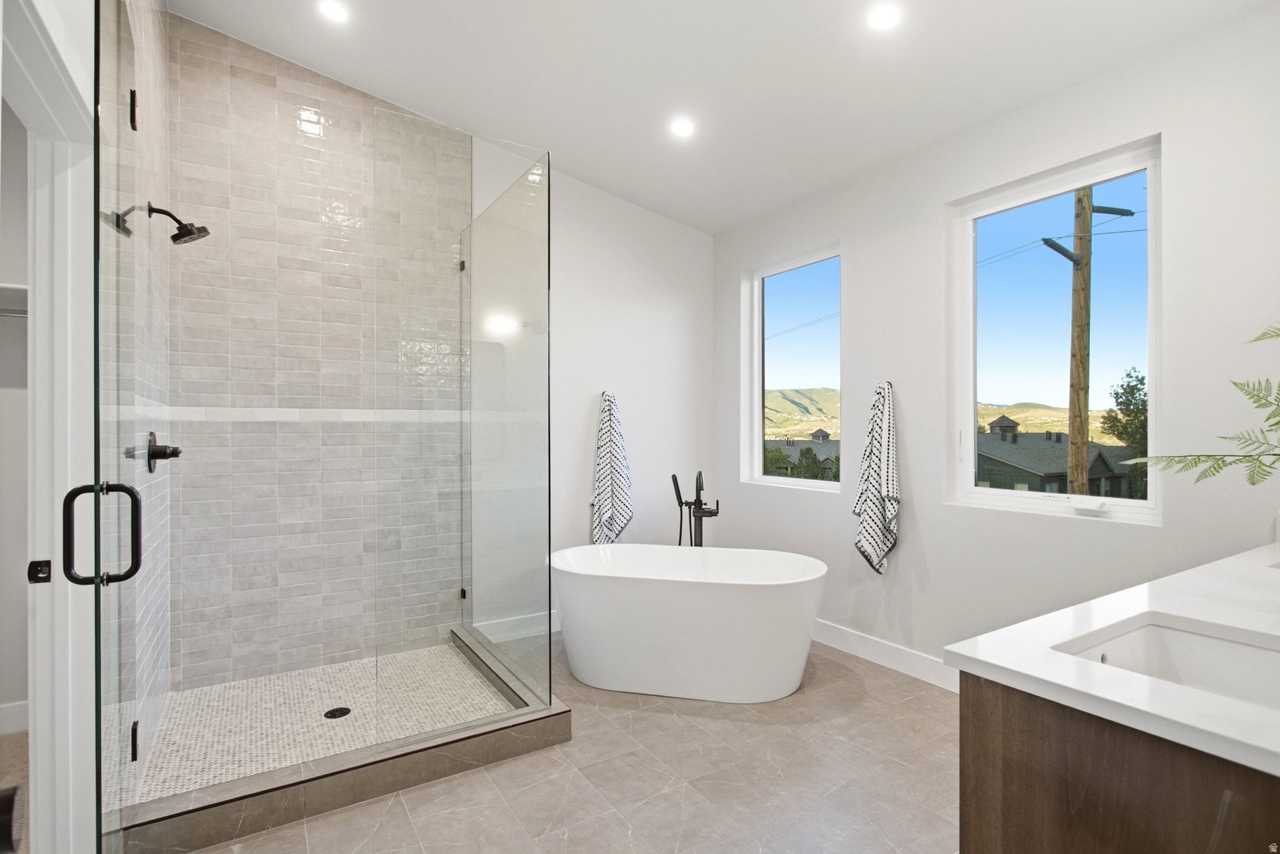 Modern bathroom with a glass shower, freestanding tub, and windows overlooking distant hills.