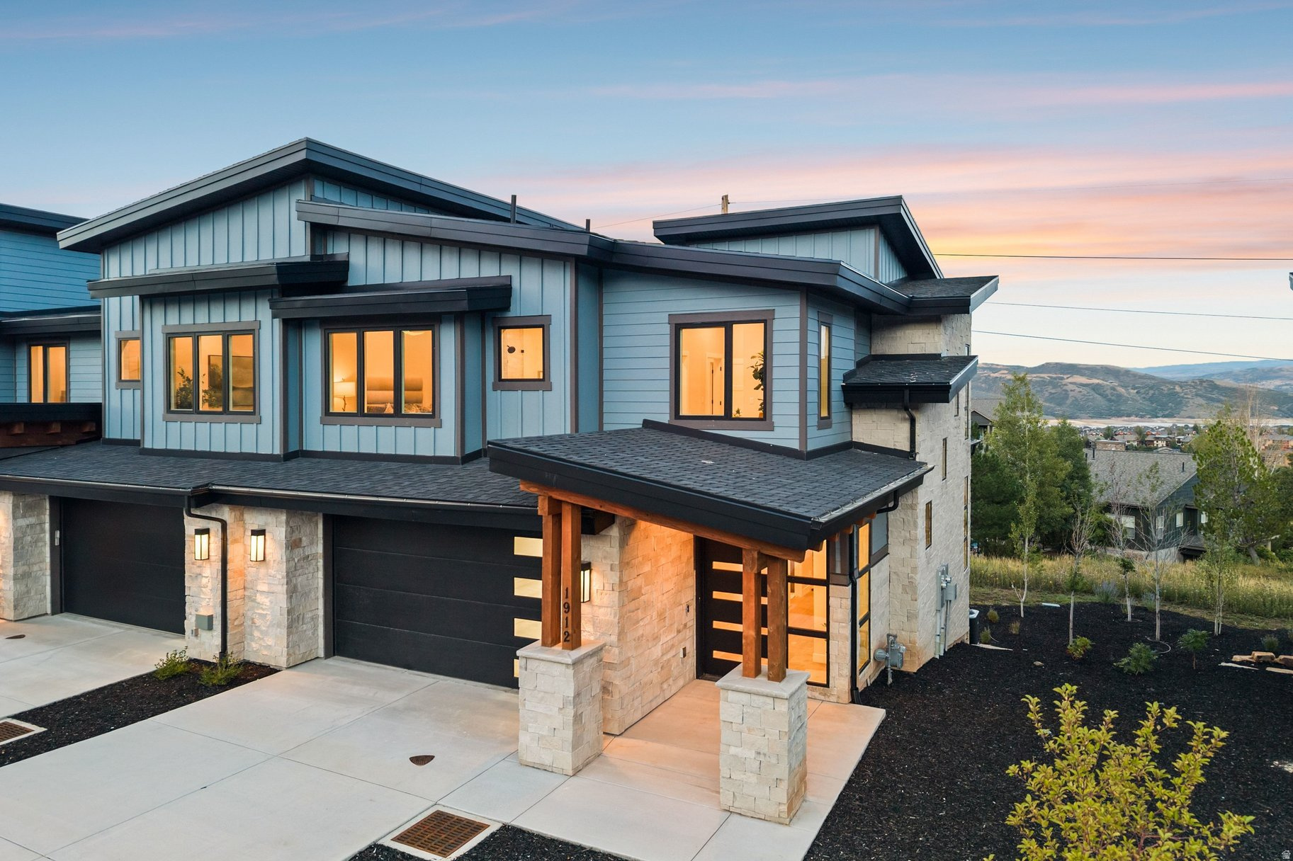 Modern blue-gray home with stone accents and a covered entry, glowing windows, and mountain views at dusk.