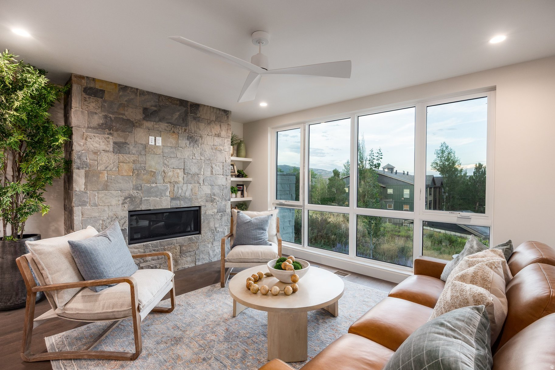 Staged living room with a stone fireplace, expansive windows, and views of trees and distant hills.