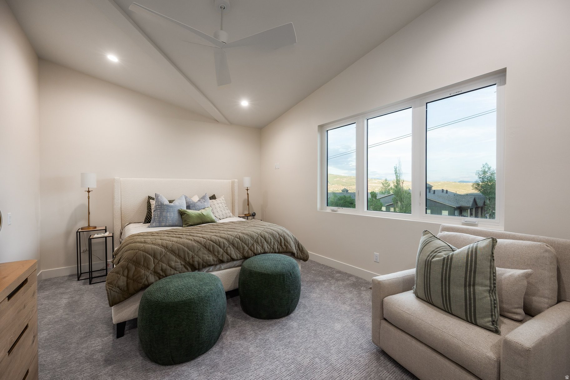 Furnished bedroom with a large bed, green ottomans, and wide windows overlooking rolling hills.