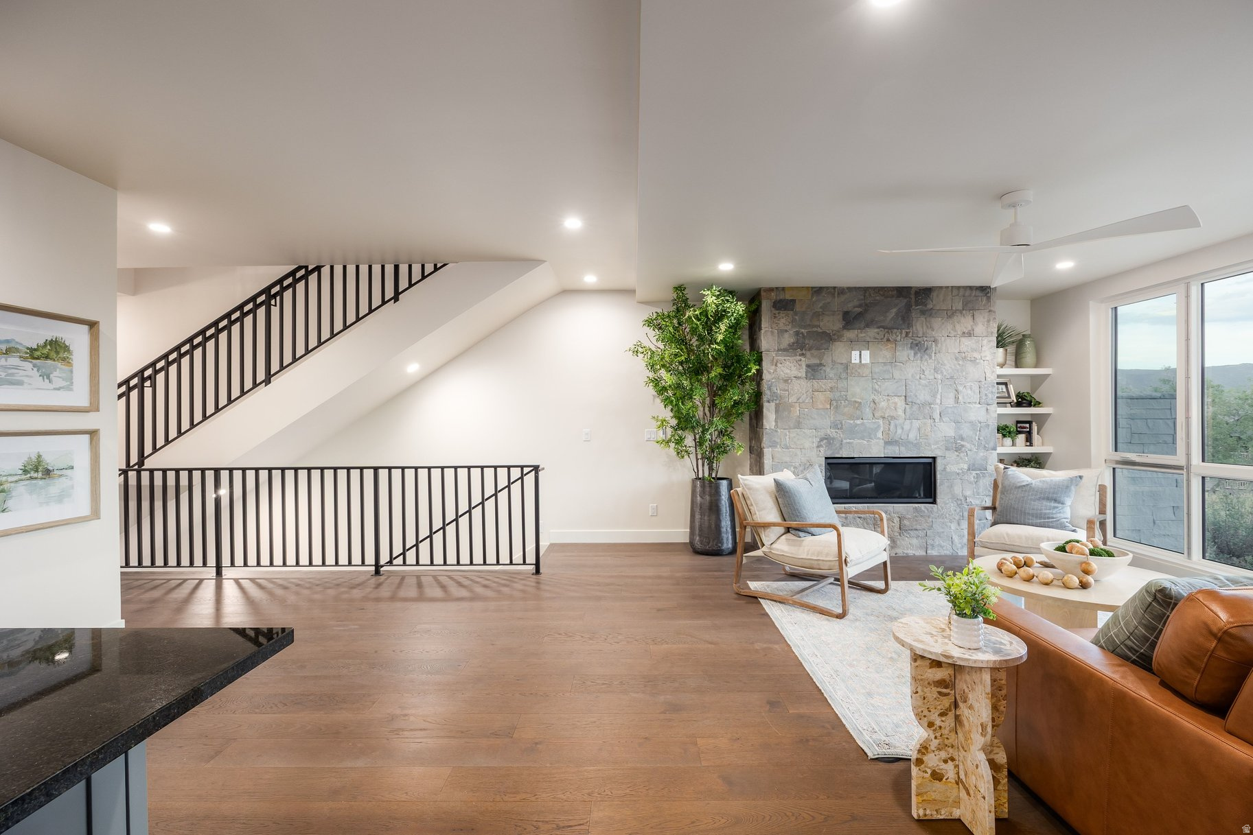 Living room with a stone fireplace, black-railed staircase, wood floors, and large windows.