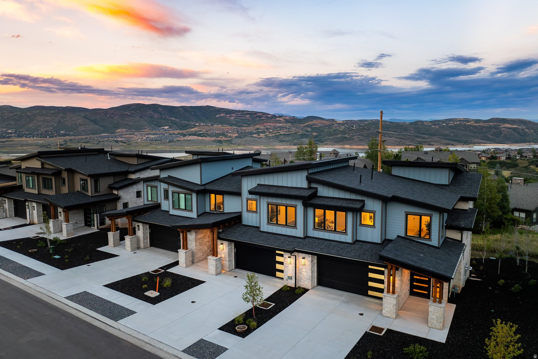 Modern homes with warm illuminated windows overlook a lake and mountain valley at dusk.