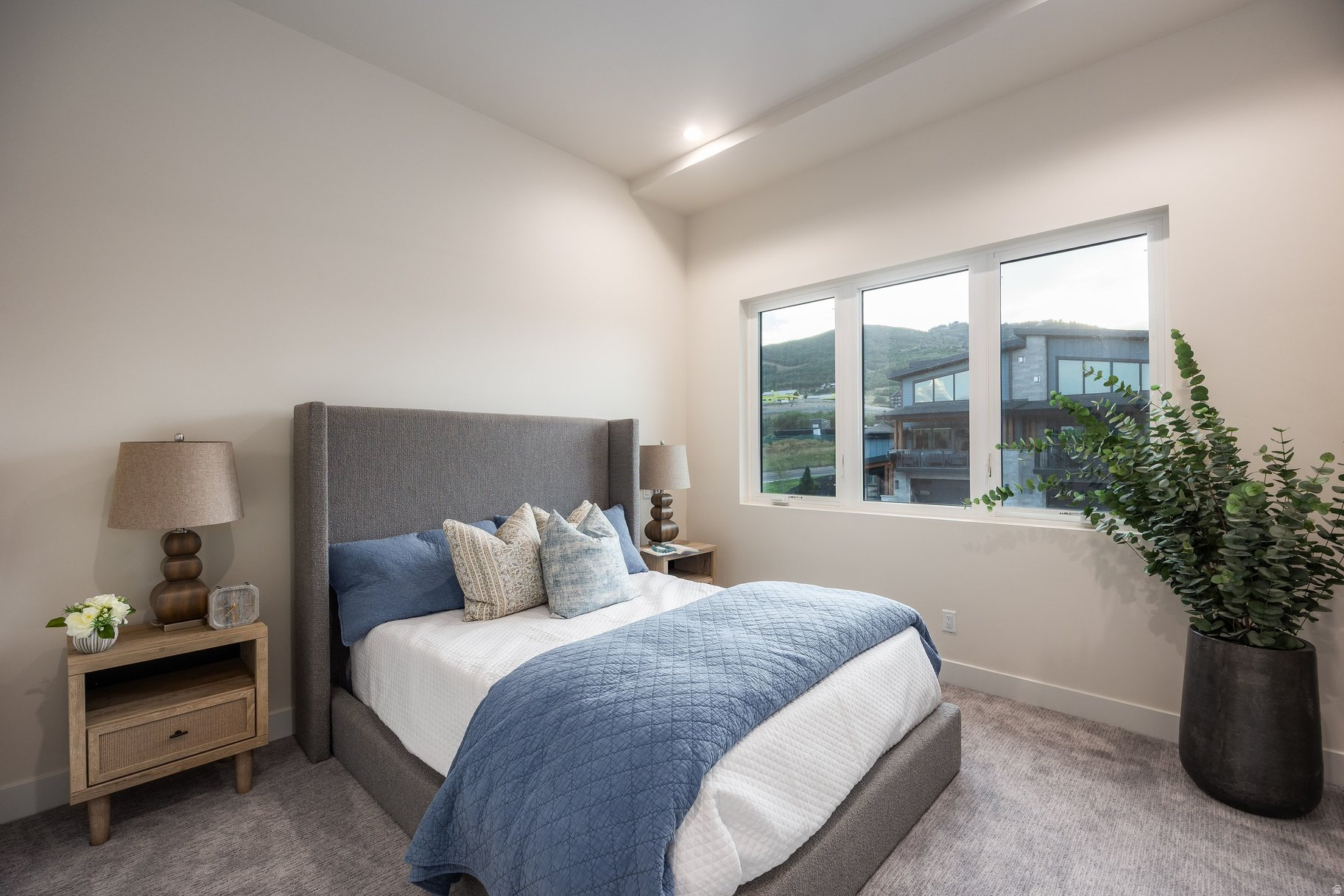 Staged bedroom with a gray upholstered bed, blue bedding, and a wide window overlooking green hills.