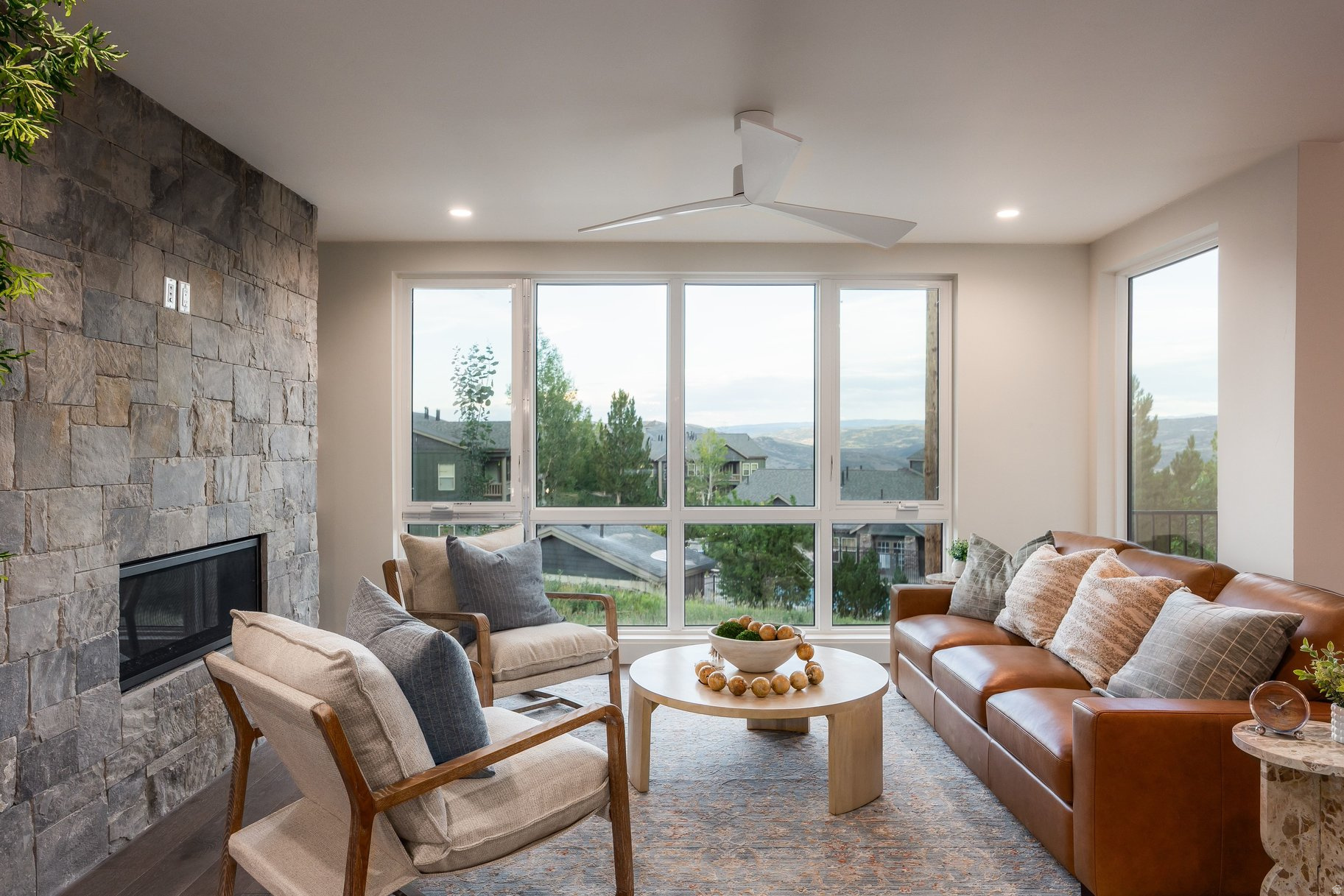 Furnished living room with a stone fireplace, leather sofa, and large windows overlooking distant hills.