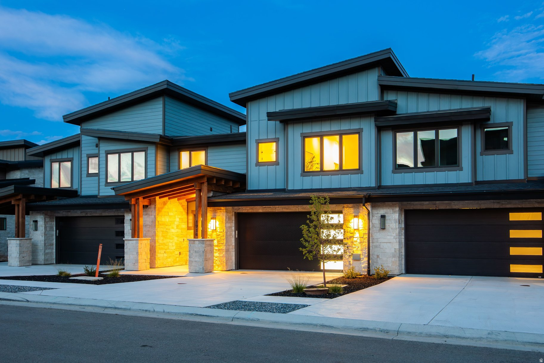 Modern blue-gray residence at dusk with a stone facade, covered entry, and warmly lit windows.