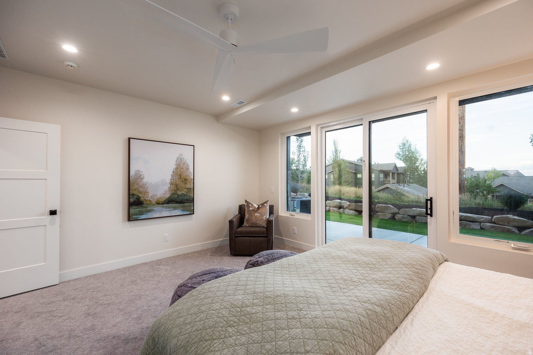 Bedroom with a pale green quilt, dark armchair, and large windows overlooking neighboring homes.