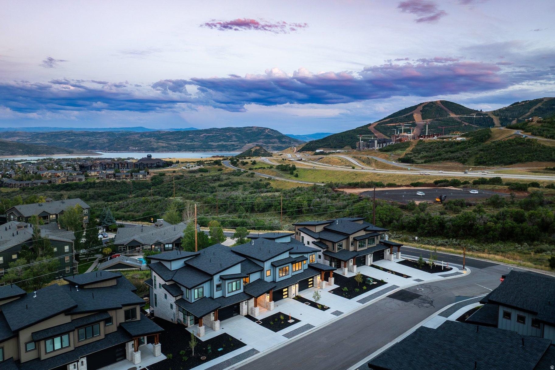 Modern homes overlook a lake and mountain ridges, with hillside construction in the distance.