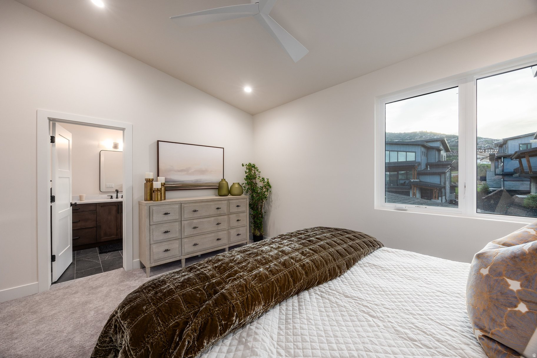 Furnished bedroom with a quilted bed, dresser, and large windows overlooking neighboring homes.