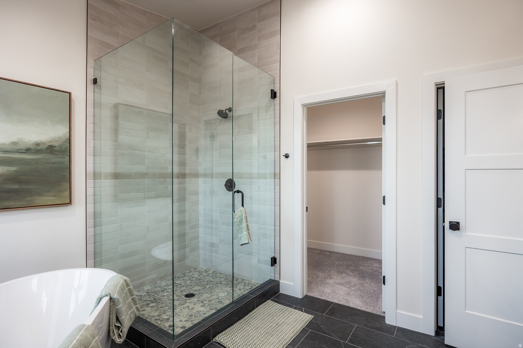 Modern bathroom with a glass walk-in shower, pebble floor, freestanding tub, and open closet doorway.