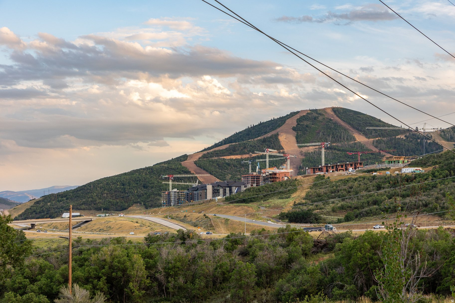 Mountain hillside with ski runs and new buildings under construction, viewed across a road and wooded foreground.