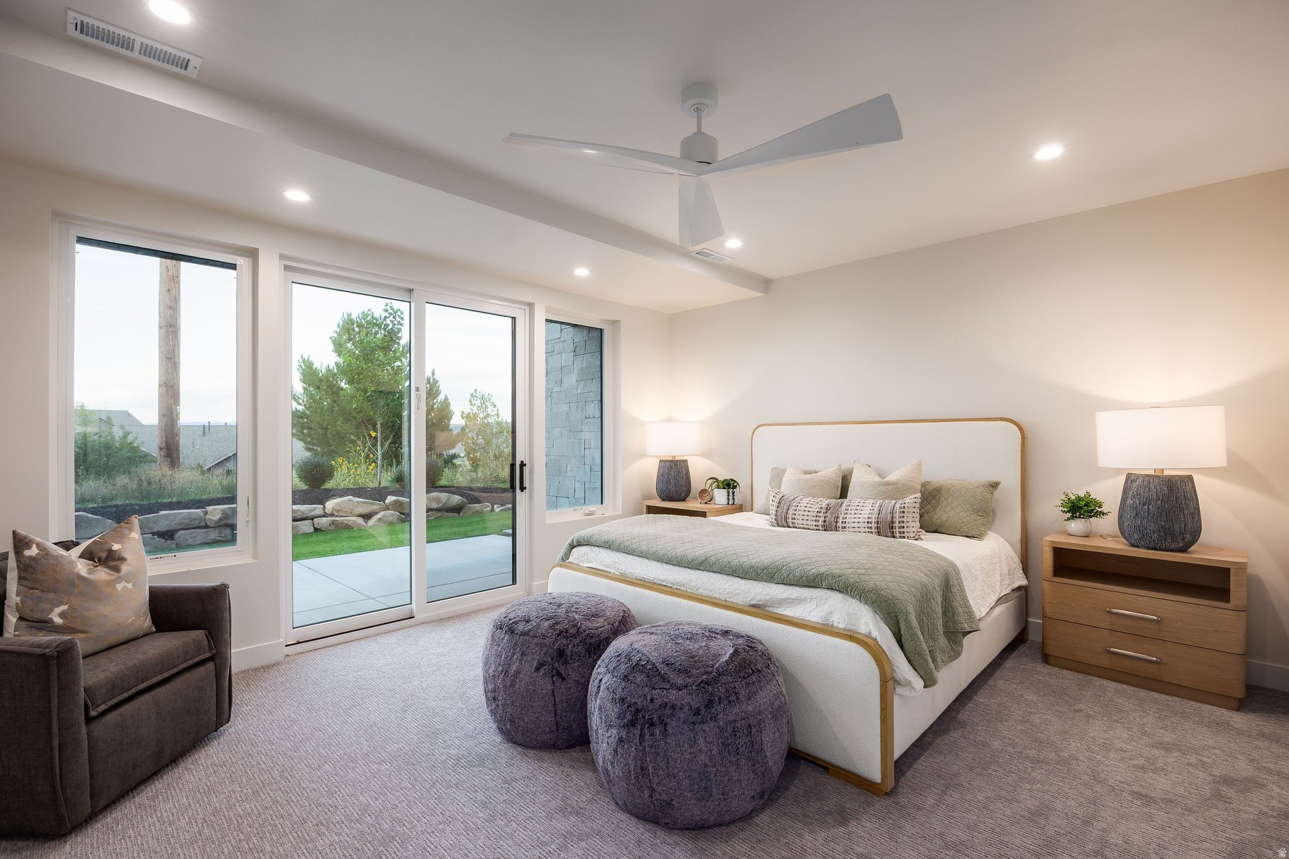 Furnished bedroom with a pale green bedspread, purple ottomans, and large windows opening to a garden.