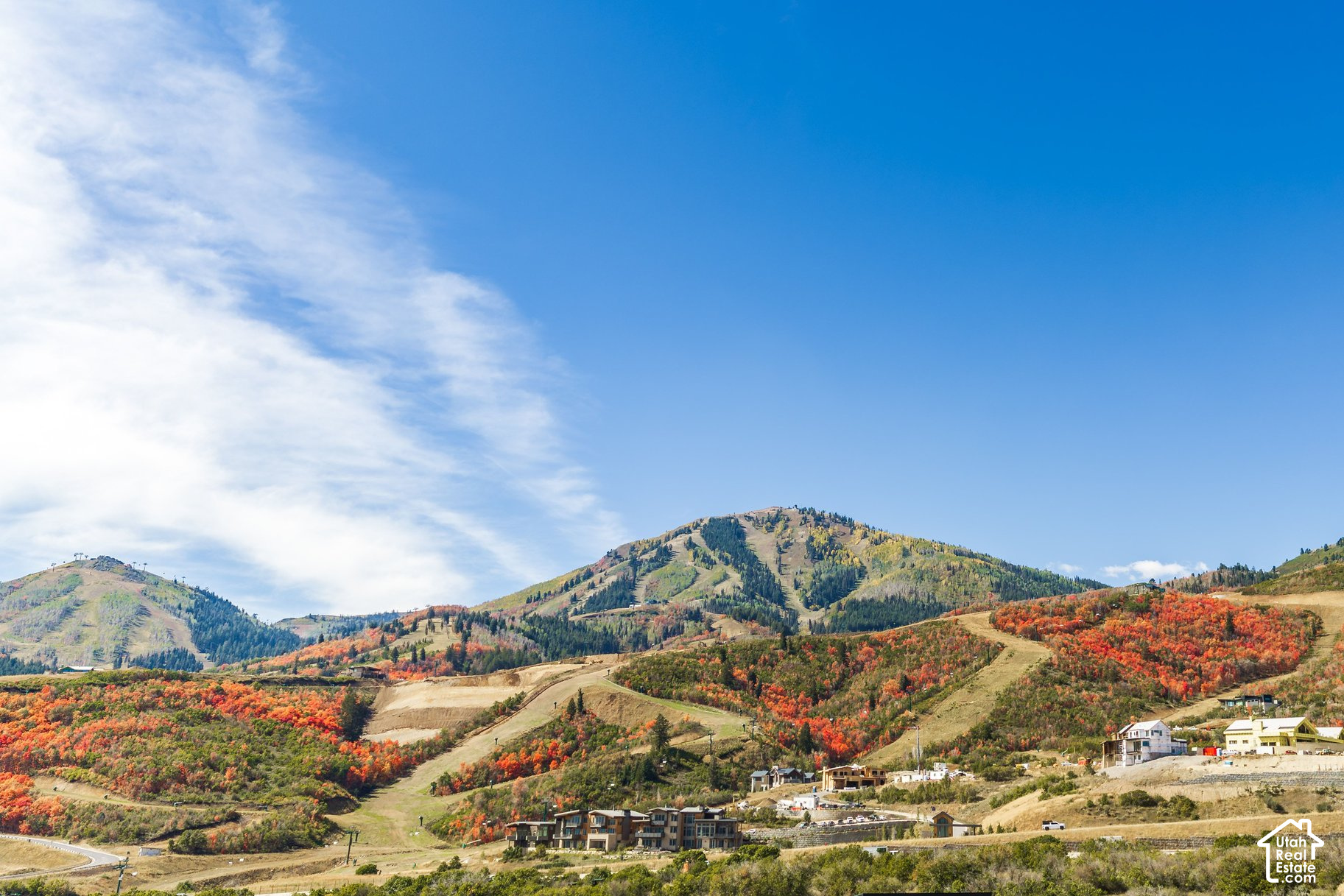 Mountain slopes with autumn foliage and ski runs above hillside homes under a blue sky.