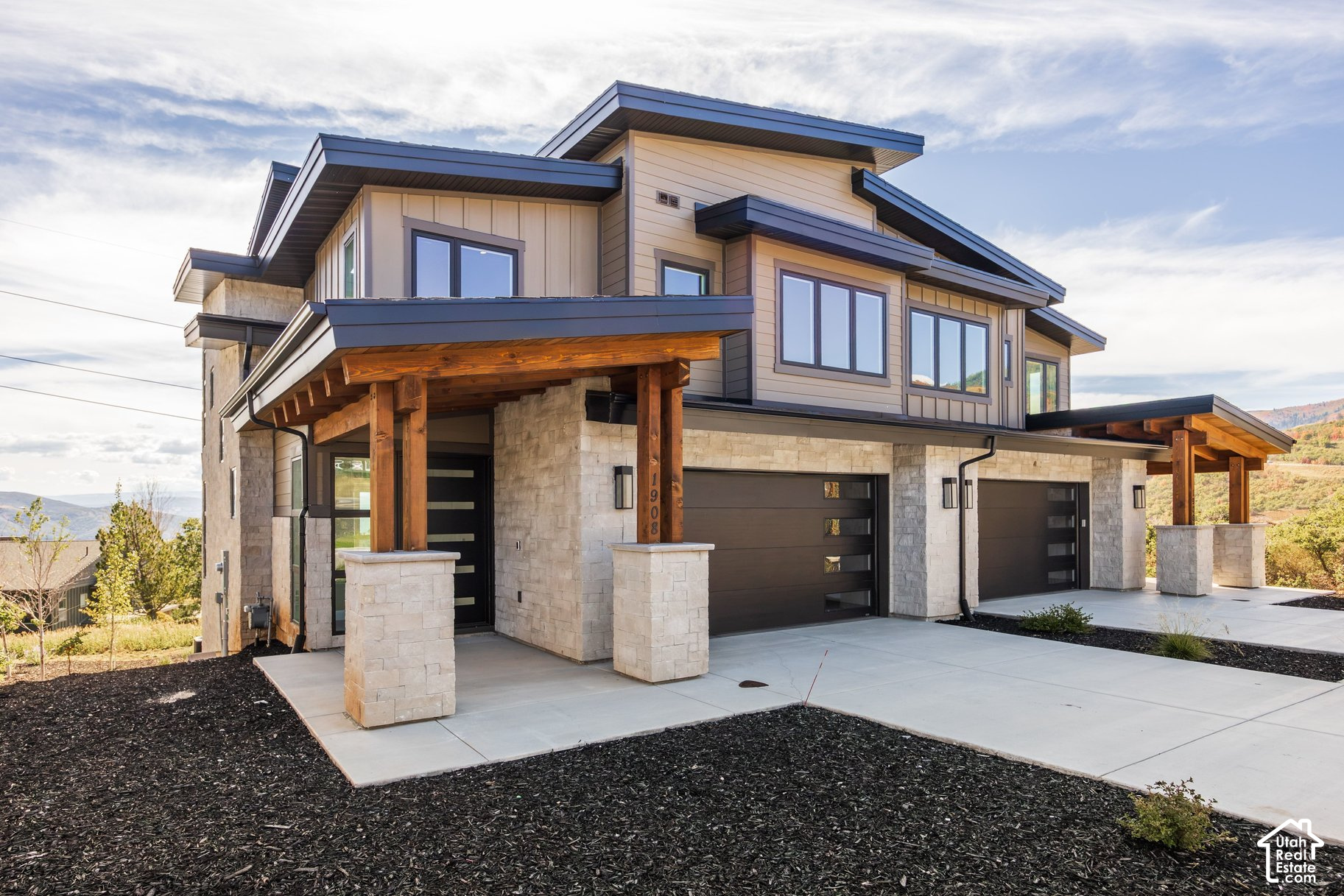 Modern two-story home with stone cladding, timber porch supports, and two garage doors on a hillside.
