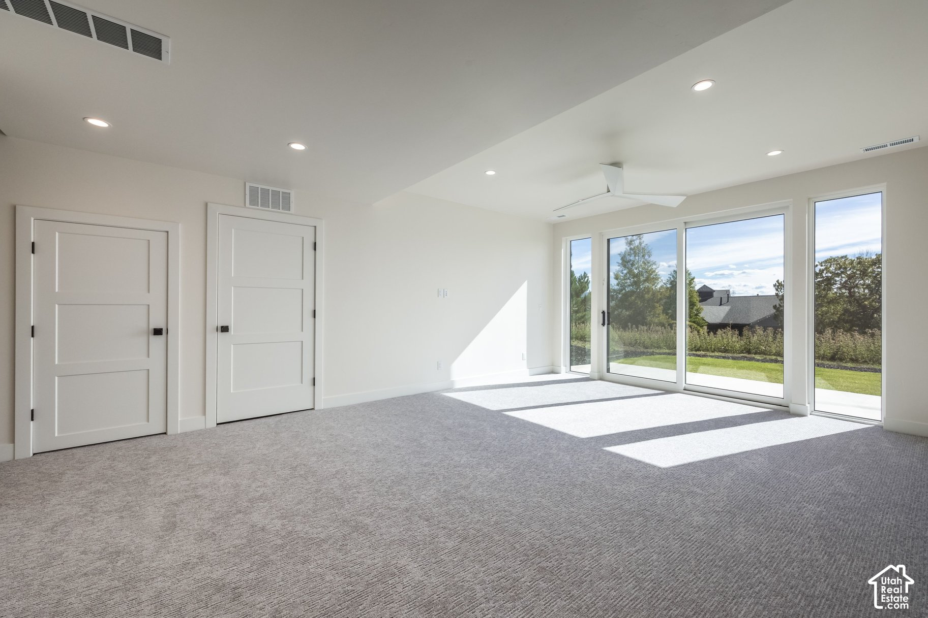 Empty carpeted bedroom with a ceiling fan and wide sliding glass doors overlooking a lawn and trees.