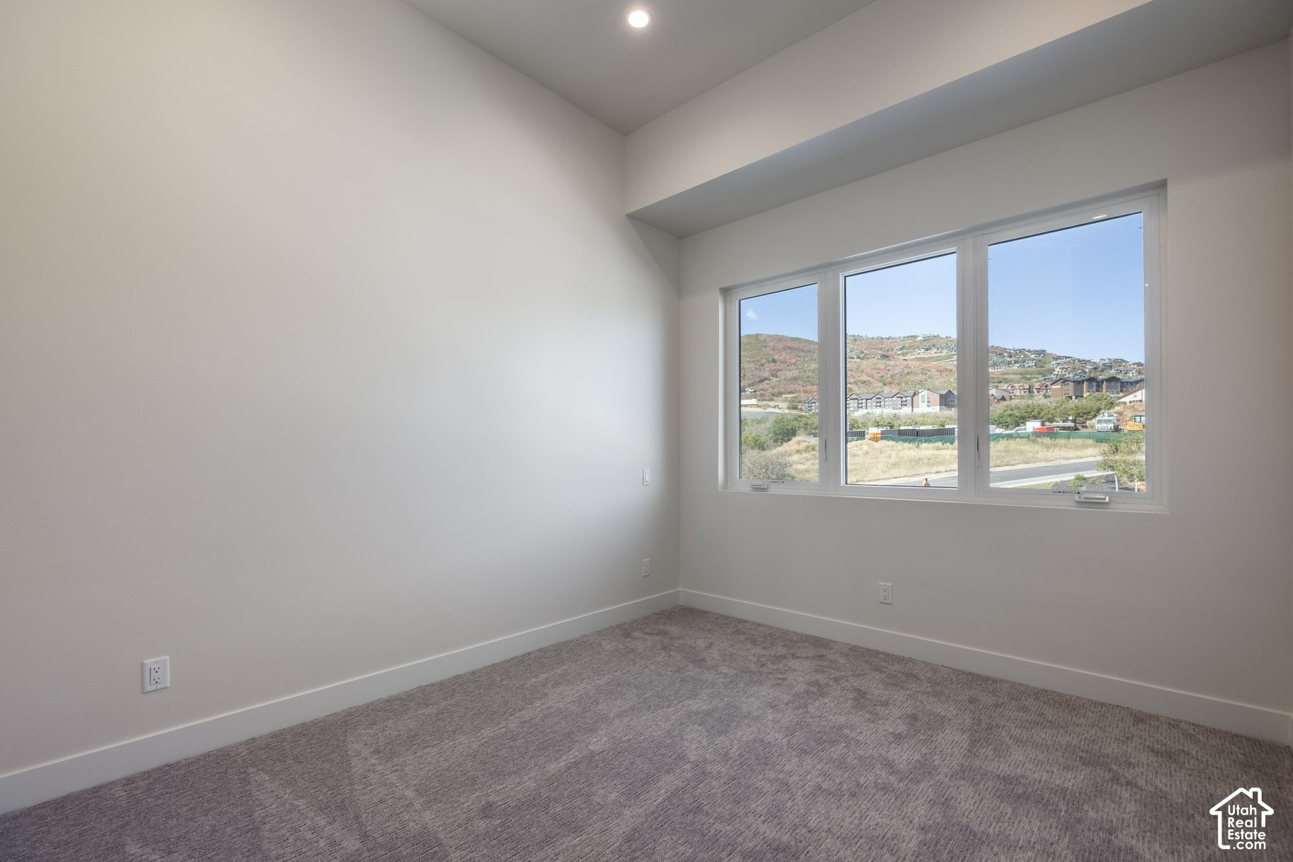 Empty bedroom with gray carpet and a wide window overlooking hillside homes.