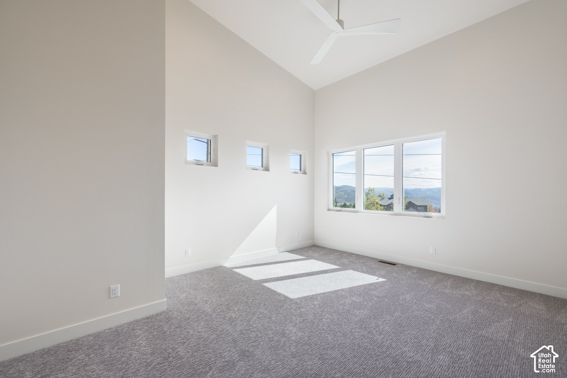 Empty carpeted room with a high ceiling, ceiling fan, and windows overlooking distant hills.