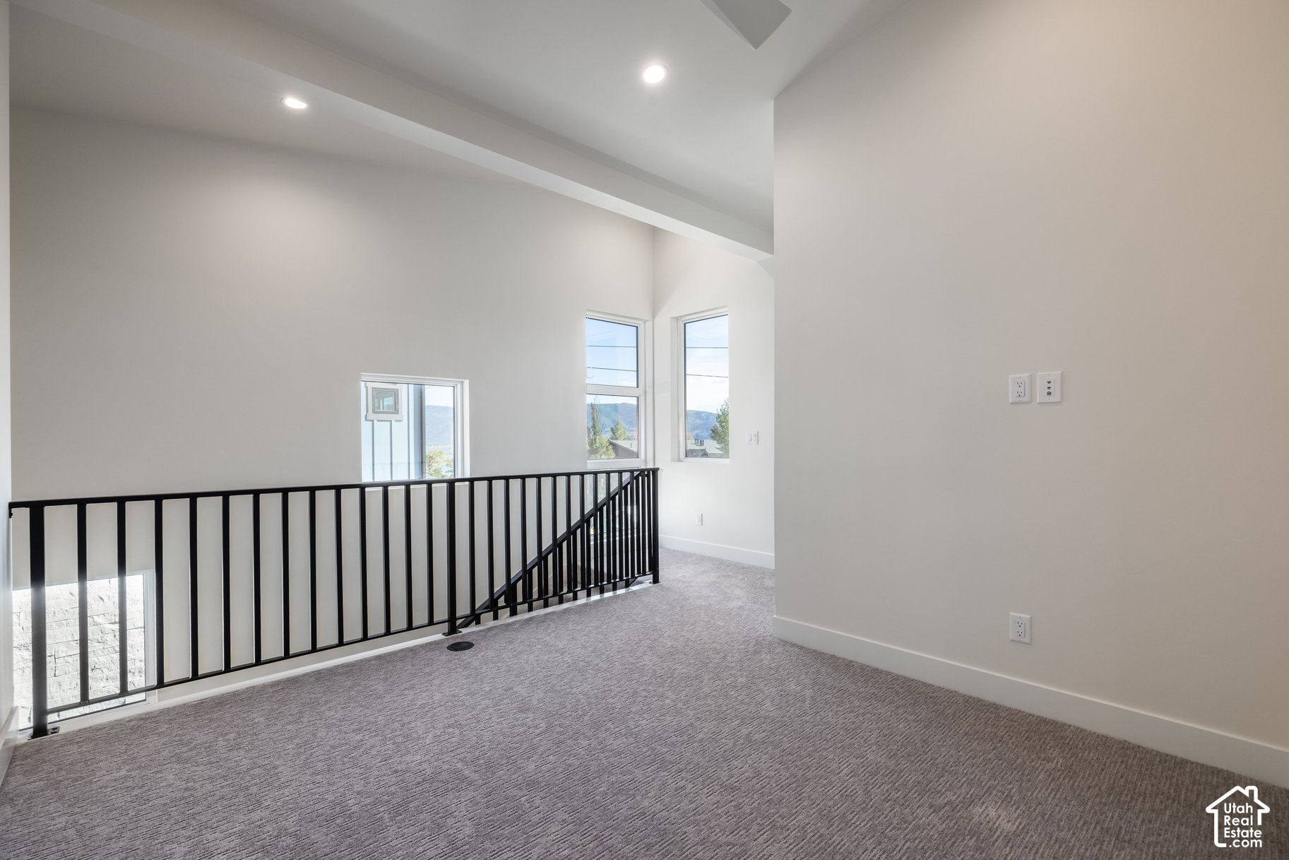 Empty carpeted upstairs loft with a black stair railing and tall windows overlooking mountains.